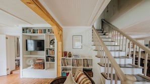 A living room with a wooden staircase, a bookshelf, a television, and a plaid sofa. - Historic farmhouse by the river in Keene, NY near Lake Placid & Whiteface Mt. (Keene)