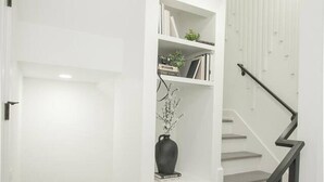 Una escalera con barandilla negra, paredes blancas y un estante con libros y un jarrón.