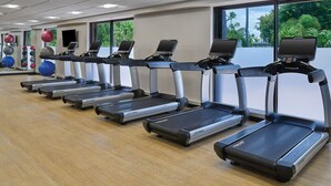 A gym with multiple treadmills aligned in a row, large windows with a view of greenery, and exercise balls in the background.