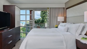 A hotel room with a large bed, a TV, a balcony with a view of the ocean, and a window with curtains.
