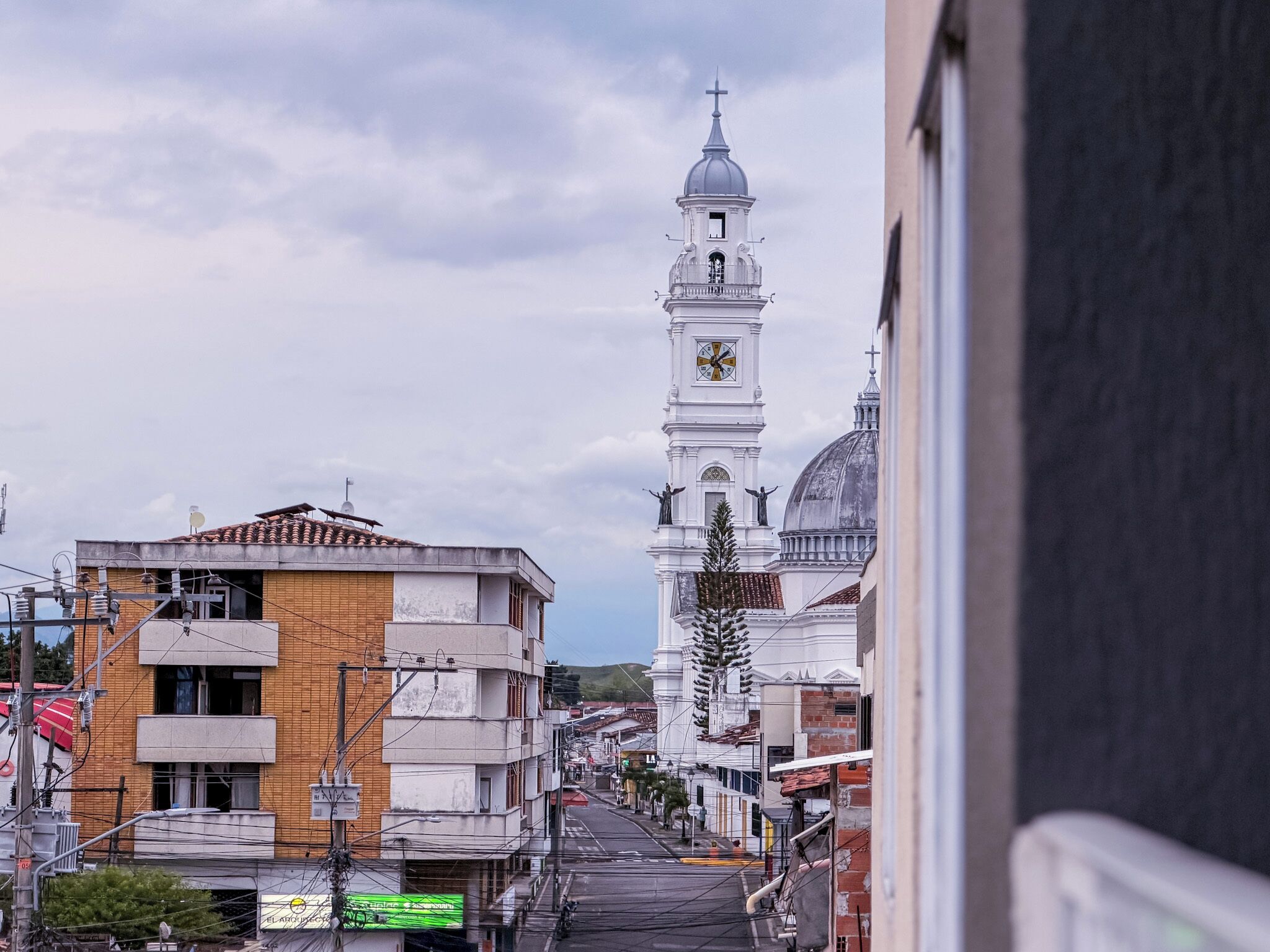 A street view with a prominent white church tower with a clock and crosses, surrounded by buildings with various architectural styles.