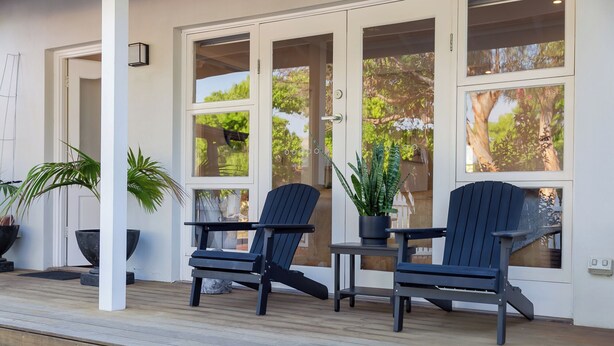 Een overdekt terras met twee Adirondack-stoelen en een kleine tafel, omgeven door grote ramen met uitzicht op bomen en een heldere lucht.