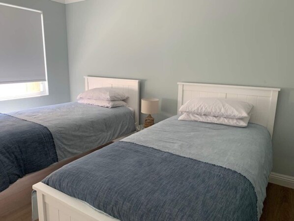 Two twin beds with white headboards and grey bedding, a bedside lamp, and a window with blinds.
