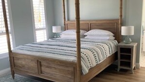 Una cama con dosel de madera y un edredón estampado, una mesita de noche con lámpara, y un ventilador de techo en una habitación con paredes azul claro y una alfombra estampada.