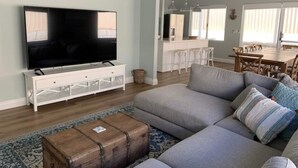 Una sala de estar con un televisor de pantalla plana grande, un sofá gris, un cofre de madera y un comedor al fondo.
