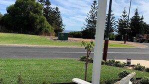 Un área de estar exterior blanca con una mesa y dos sillones, un árbol pequeño, y una vista a una calle y follaje en el fondo.