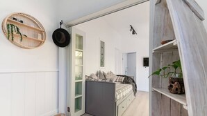 Een knus slaapkamer met een bed, een klein nachtkastje, een wandplank met een hoed, en een deur naar een andere kamer.