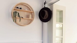 Een kamer met een witte paneelwand, een rond geweven schap met een plant, een zwarte hoed die aan de muur hangt, een rieten mand op de vloer, en een deur die leidt naar een balkon met uitzicht.