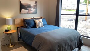 A bedroom with a large bed, a bedside table with a lamp, a painting on the wall, and a window with a view of the outside. - Modern 1-bedroom cottage in historic downtown Mount Vernon. (Mount Vernon)