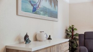 Una habitación con un mostrador blanco, un cuadro enmarcado de una hamaca sobre el agua, un jarrón blanco y una pequeña figurita de foca.