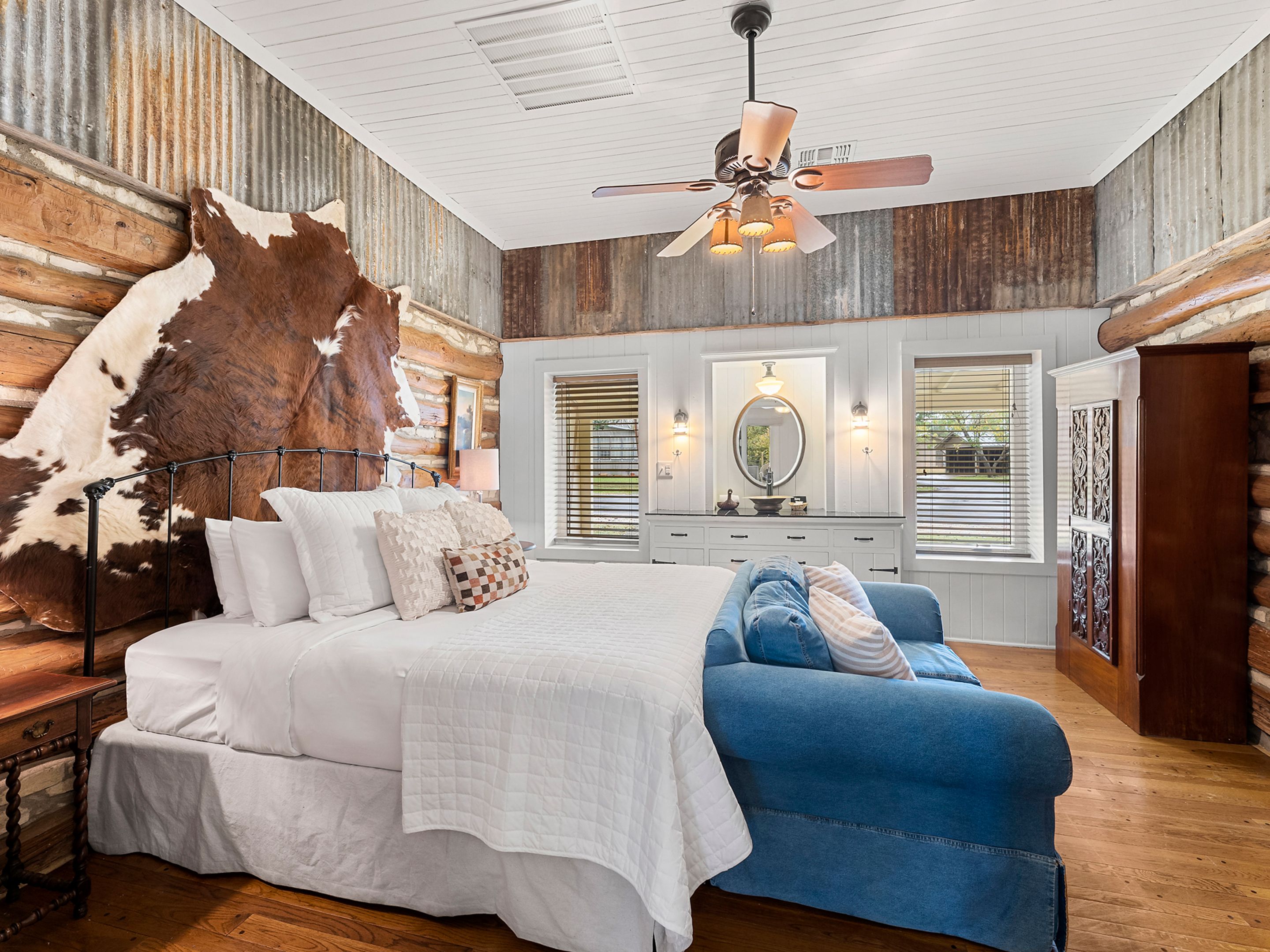 A bedroom with a large bed, a blue sofa, a wooden dresser, a mirror, and a ceiling fan.
