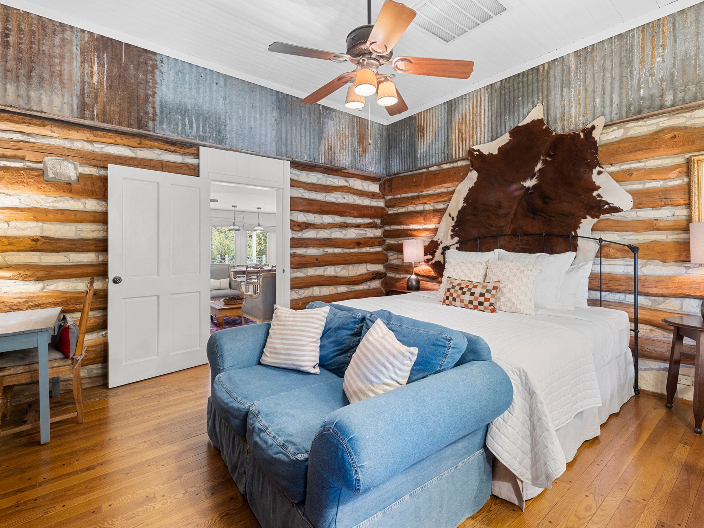 A log cabin-style bedroom with a bed, a blue couch, a ceiling fan, and a rustic wooden wall.
