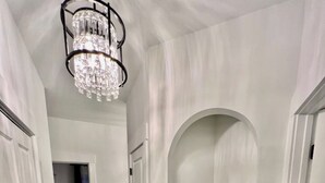 Un pasillo con paredes blancas, piso de madera y una araña. A la izquierda hay una puerta y a la derecha un estante con objetos decorativos.