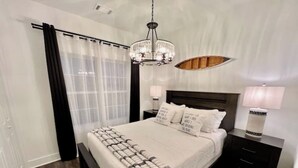 Un dormitorio con una cama grande, cabecera de madera, candelabro, mesita de noche con lámpara y ventana con cortinas traslúcidas.