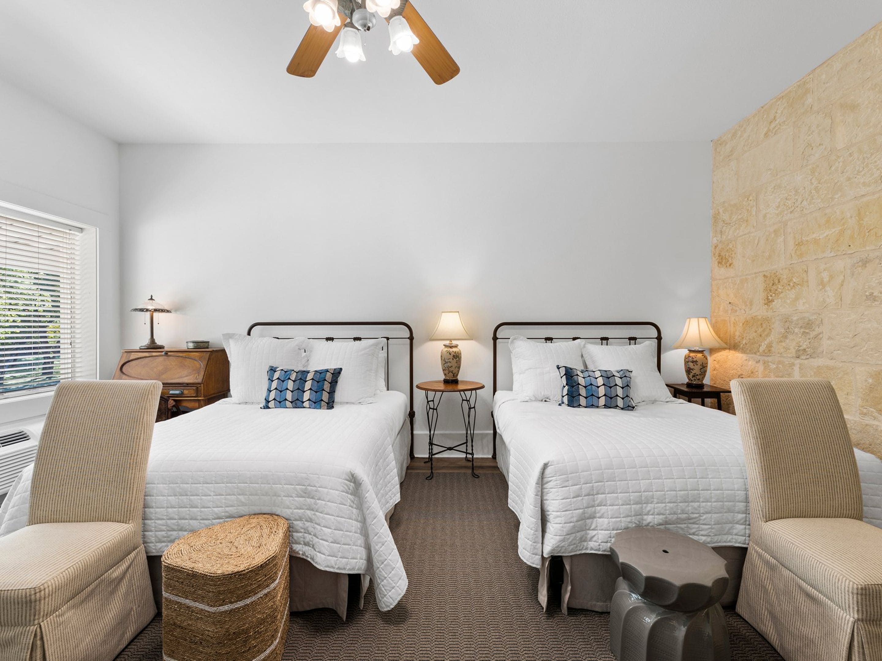 A hotel room with two beds, a ceiling fan, bedside lamps, a small table, and a stone accent wall.