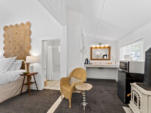 A modern hotel room with a large bed, a wicker chair, a small round table, a bathroom with a mirror, sink, and a stove.