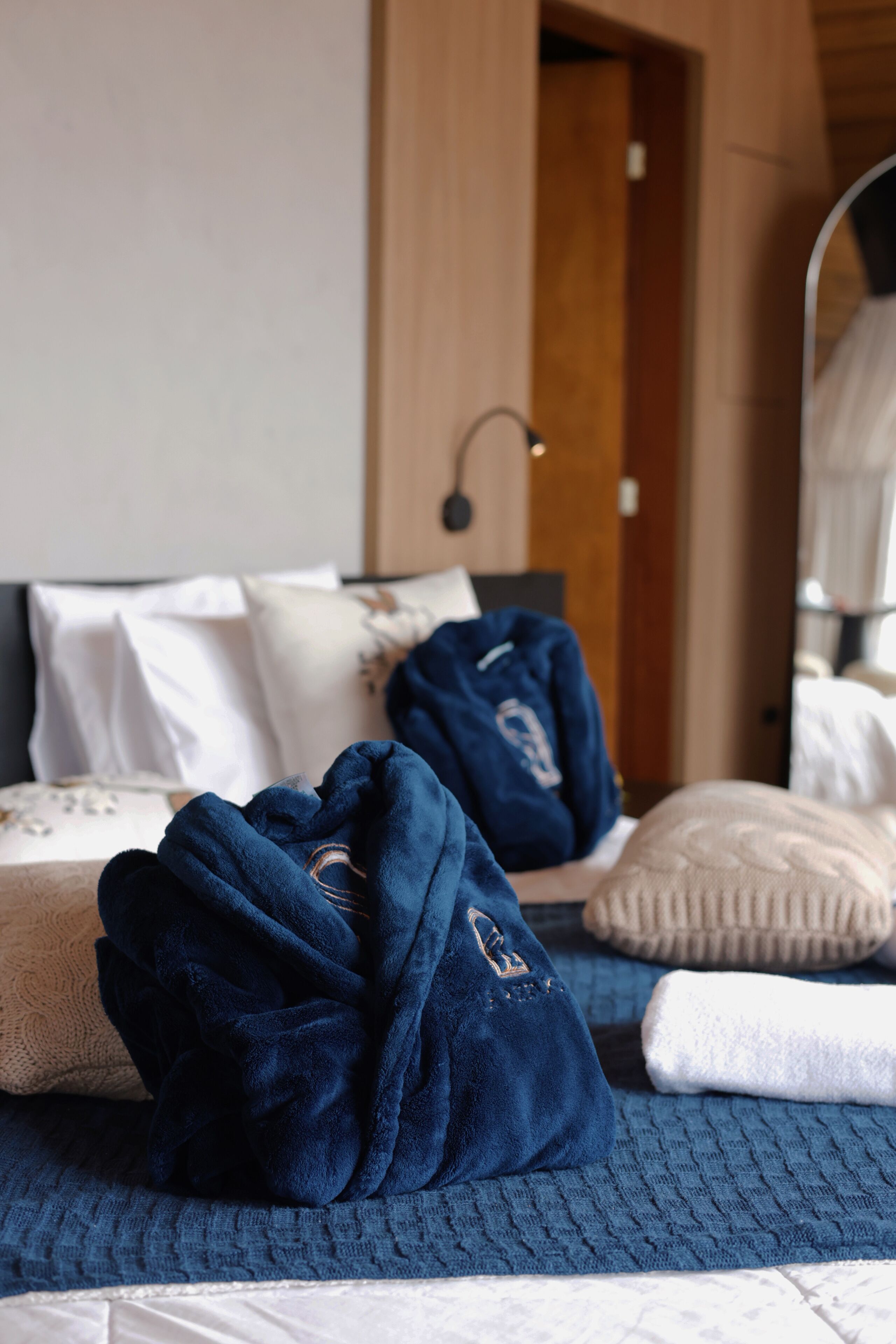 A neatly made bed with a blue blanket and pillows, a bedside table with a lamp, and a wooden door in the background.