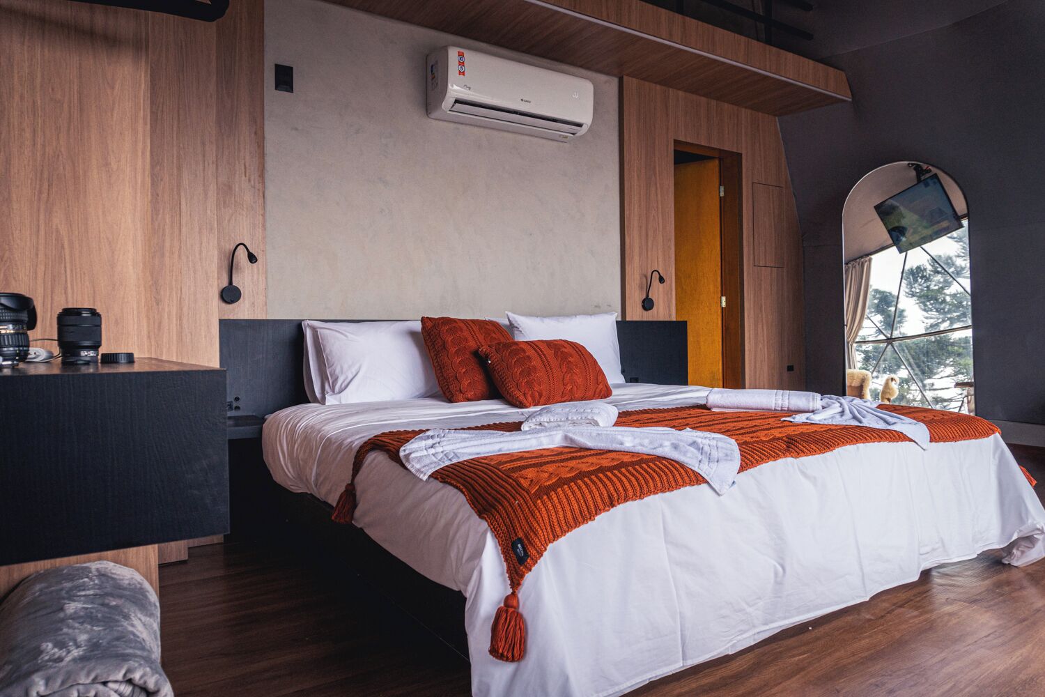 A modern hotel room with a wooden floor, a bed with white and orange bedding, an air conditioning unit, and a window with a view of trees.