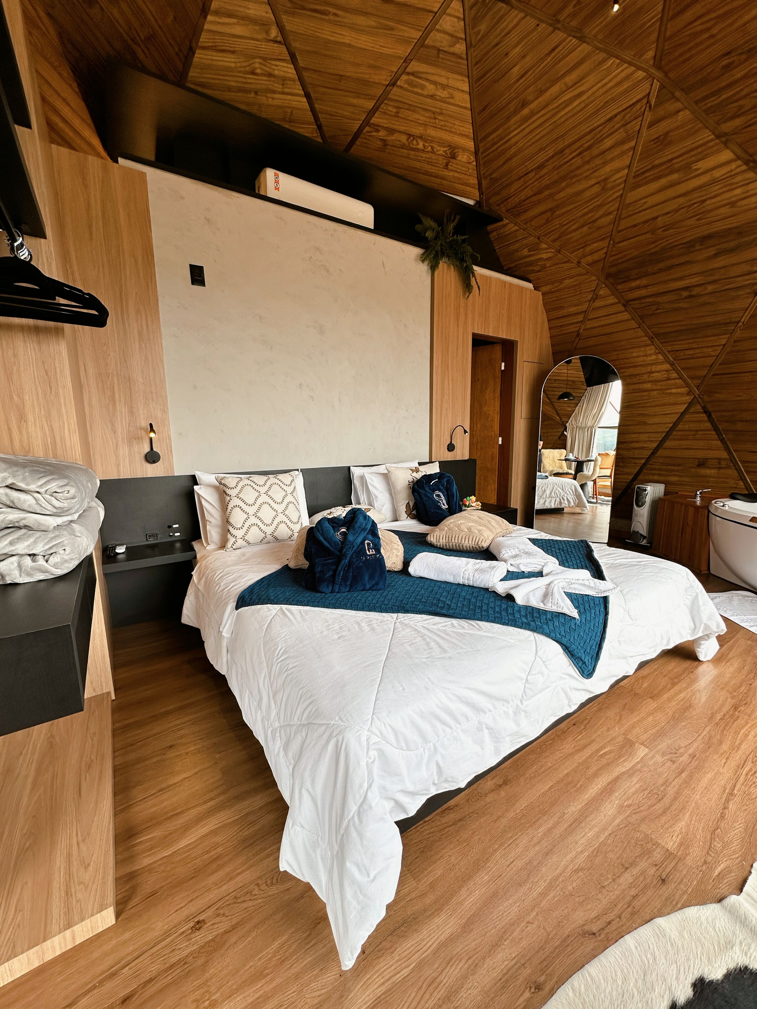 A modern bedroom with a wooden ceiling, a bed with white and blue bedding, a bedside table, and a mirror.