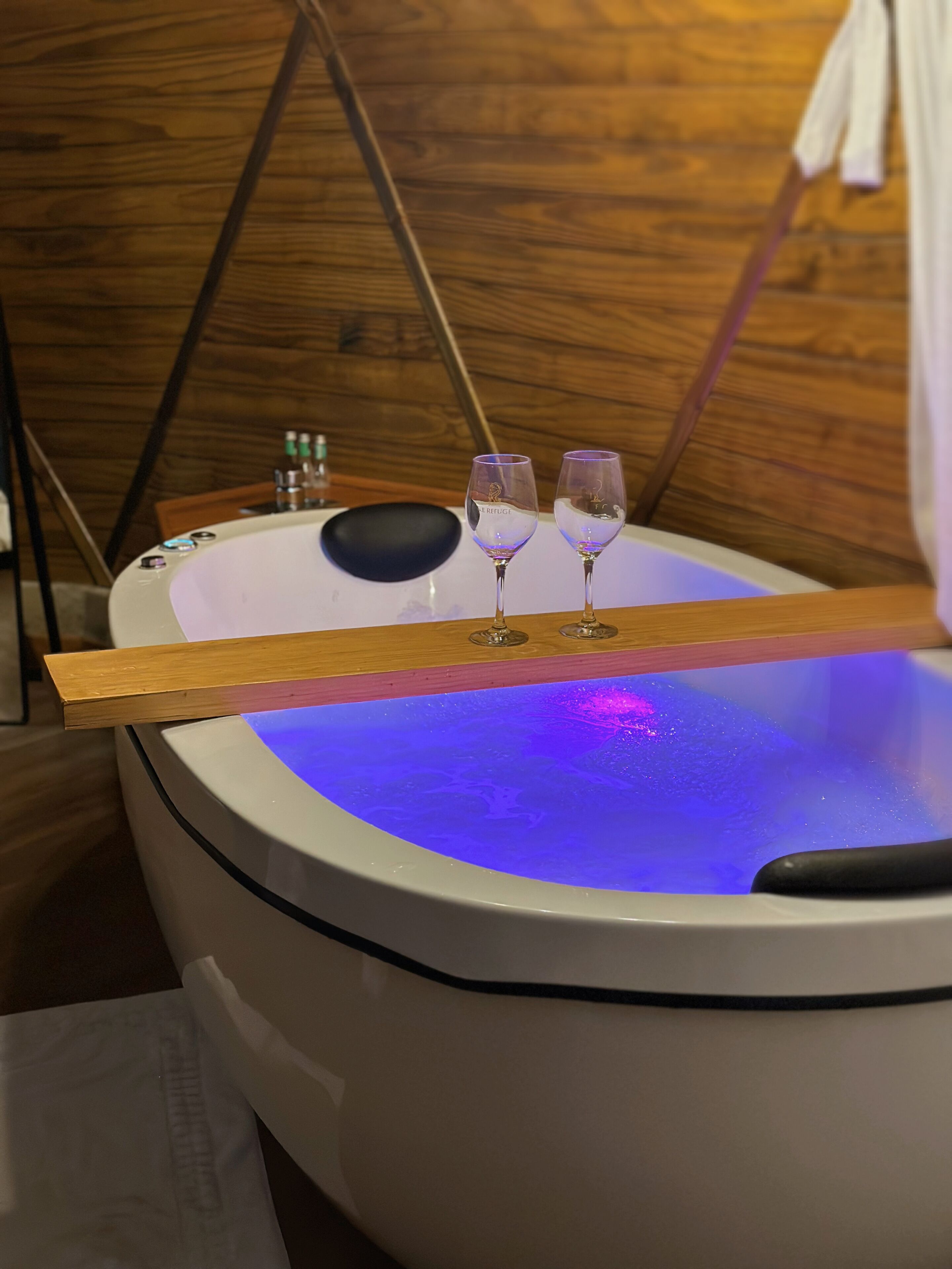 A bathtub with a wooden tray and two glasses, set against a wooden wall.
