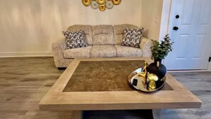 A living room with a beige sofa, patterned cushions, a coffee table with decorative items, and a wall art piece. - Luxury City Loft in Gainesville B (Gainesville)