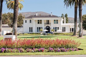 A large, two-story building with a symmetrical facade, a central entrance with a black arch, and a well-maintained lawn with colorful flower beds in the foreground.
