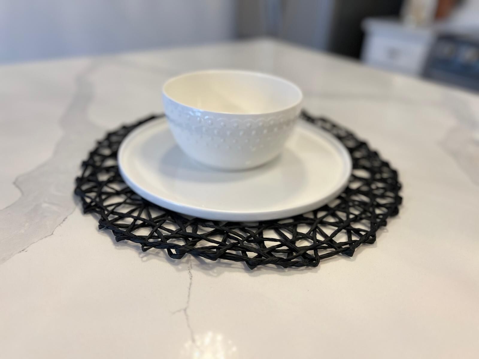 A white ceramic cup and saucer on a white table with a black woven placemat.