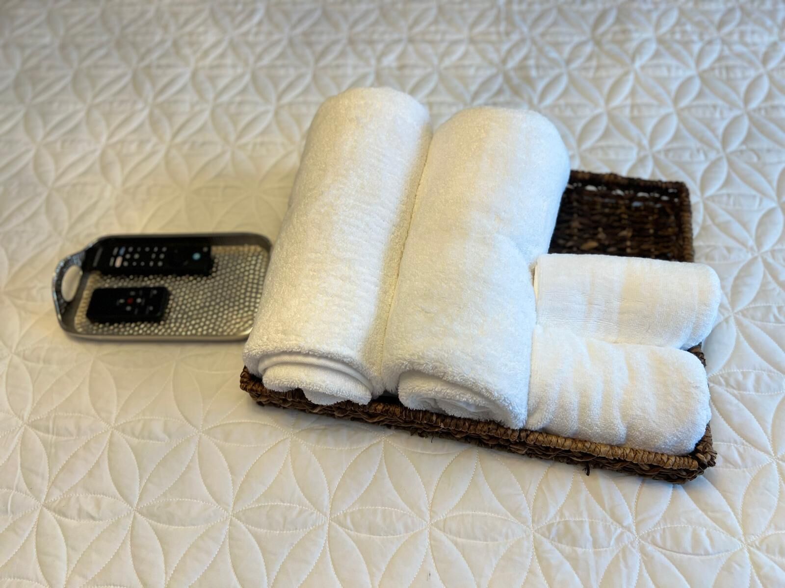Una cama con una cubierta blanca acolchada, dos toallas blancas impecables dobladas y un control remoto negro sobre la cama.