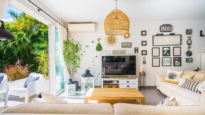 Ett modernt vardagsrum med en tv, en soffa och ett soffbord. Det finns växter, en lampa av rotting och inramade tavlor på väggen.