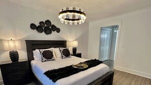 Un dormitorio moderno con una cama grande, cabecera de madera oscura, dos mesitas de noche con lámparas, una araña y un espejo en la pared.