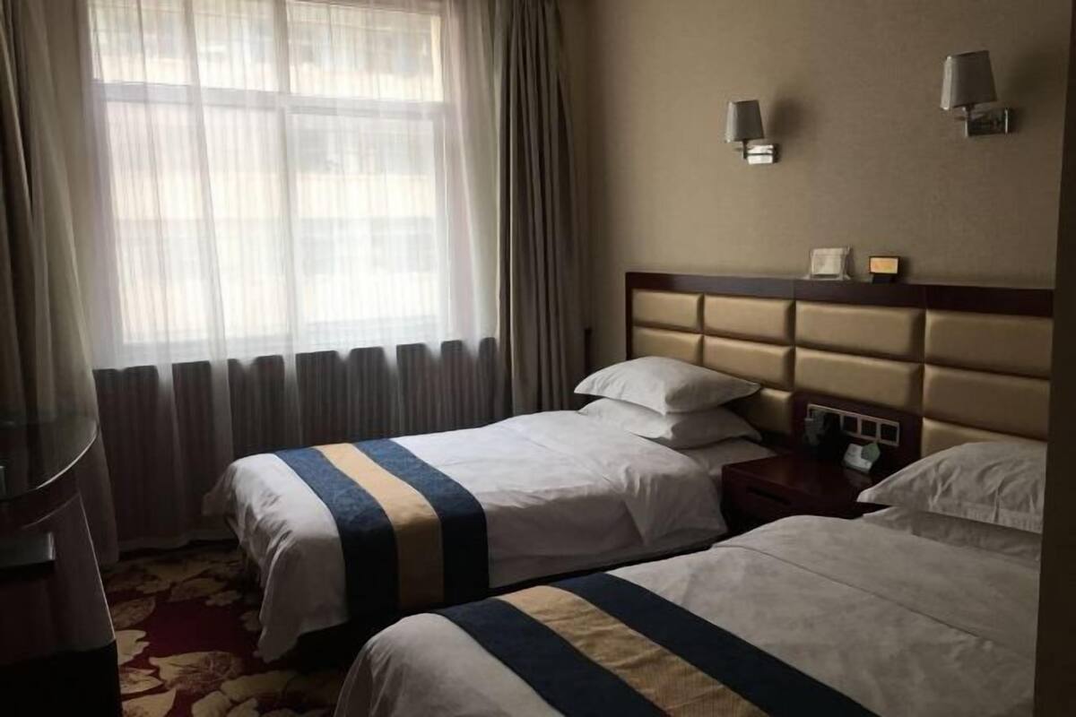 A hotel room with two beds, a nightstand, a lamp, and a window with curtains.