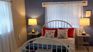Une chambre à coucher comprenant un lit, deux tables de chevet, une commode, une fenêtre avec des rideaux et un petit tabouret.