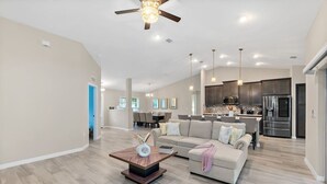 A spacious living area with a modern kitchen in the background, a dining area, and a large window providing natural light.