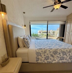 Een slaapkamer met een bed, een plafondventilator, een nachtkastje en uitzicht op zee door een groot raam.
