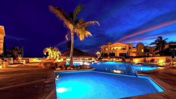 A nighttime resort pool area with a palm tree, lounge chairs, and a building with a thatched roof. - Remodeled 2025 Beachfront Escape – Pool, Views & Walkability (Cabo San Lucas)