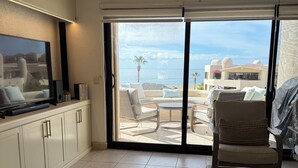 A modern living room with a flat-screen TV, a sofa, a chair, and a view of the beach through large sliding doors. - Remodeled 2025 Beachfront Escape – Pool, Views & Walkability (Cabo San Lucas)