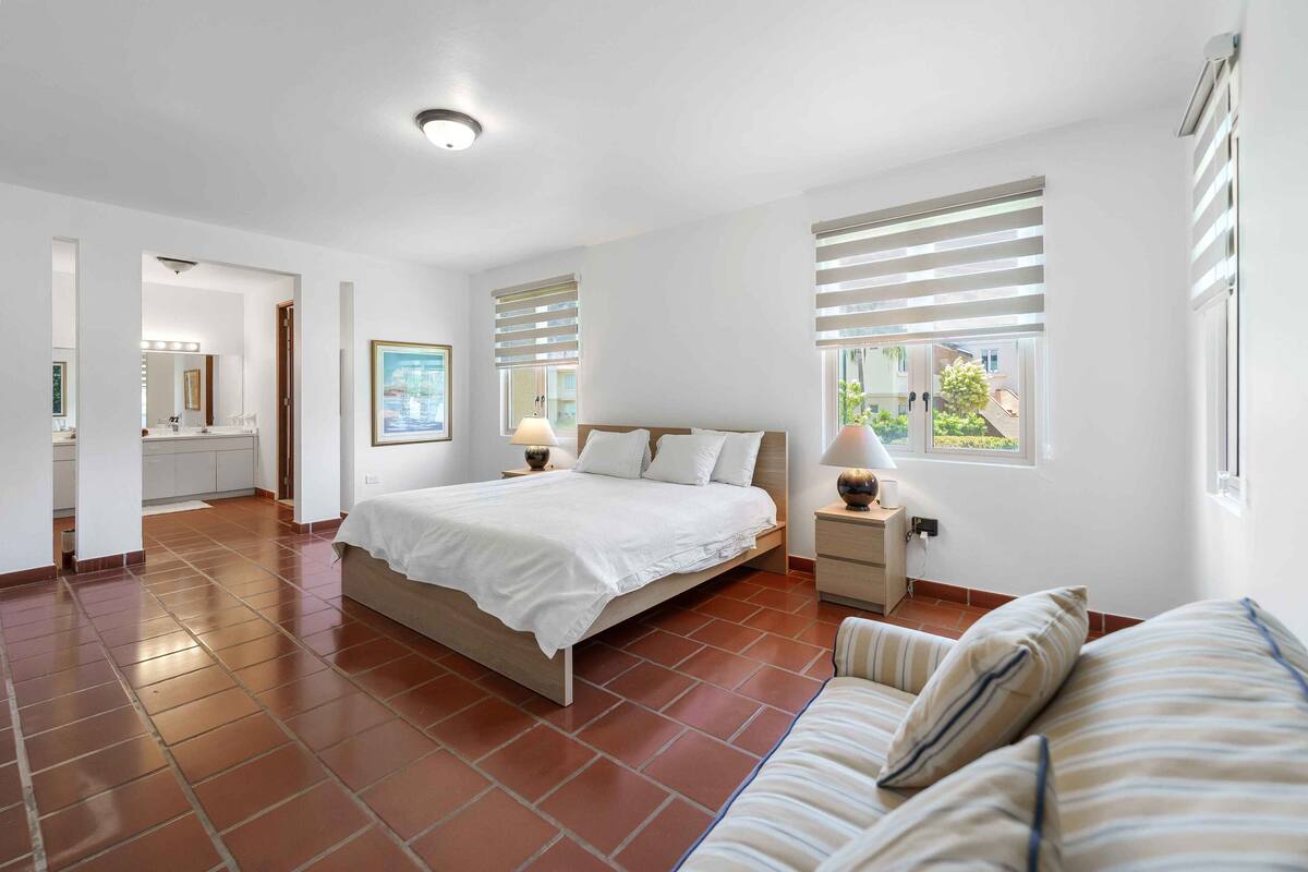 A bedroom with a bed, bedside tables, a nightstand, a window with blinds, and a view of the outside.