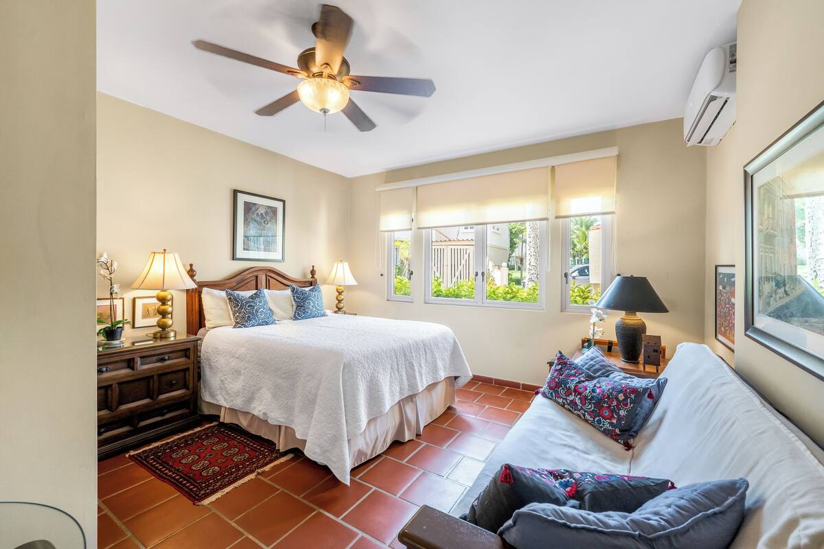 A bedroom with a bed, a sofa, a ceiling fan, a window, and a nightstand with lamps.