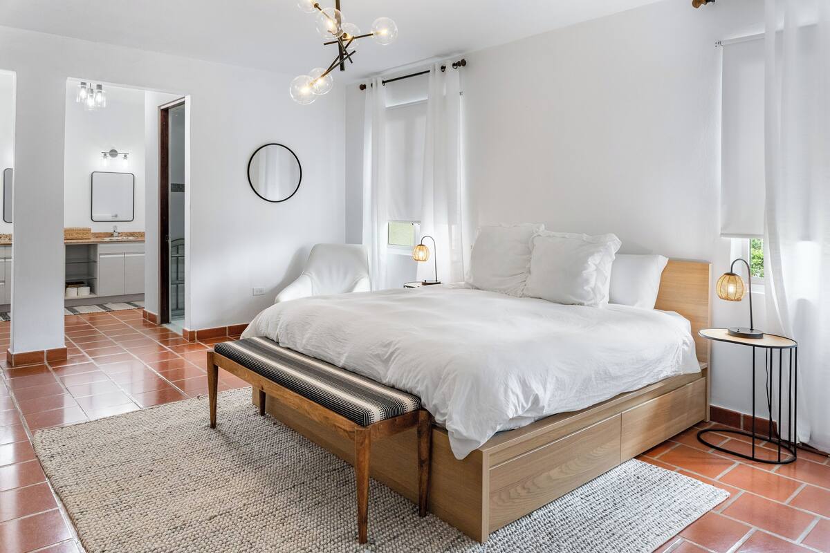 A bedroom with a wooden bed, white bedding, a bench, a bedside table with a lamp, and a view of the bathroom.