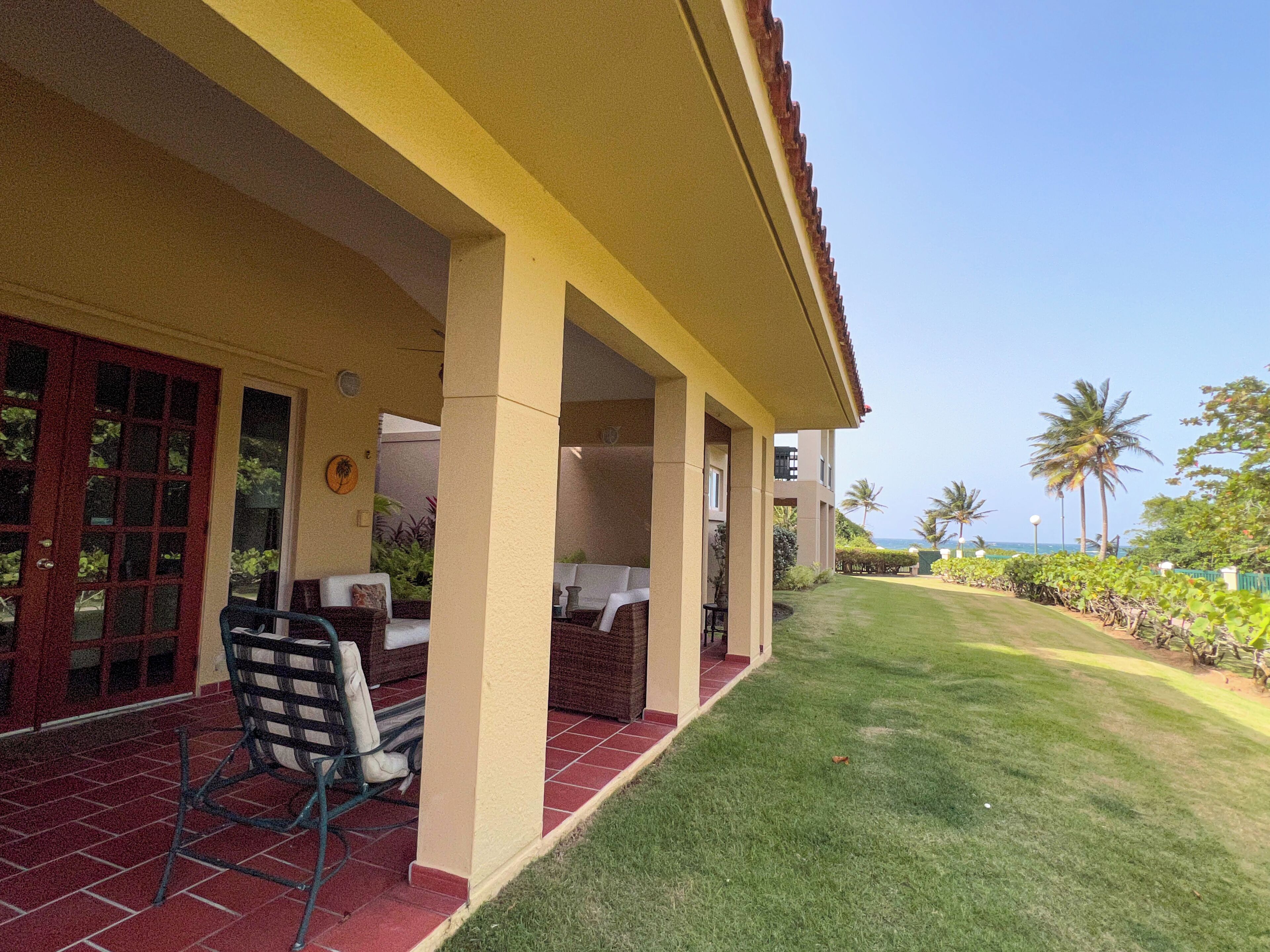 A covered outdoor area with a dining table and chairs, a red door, and a view of the ocean with palm trees.