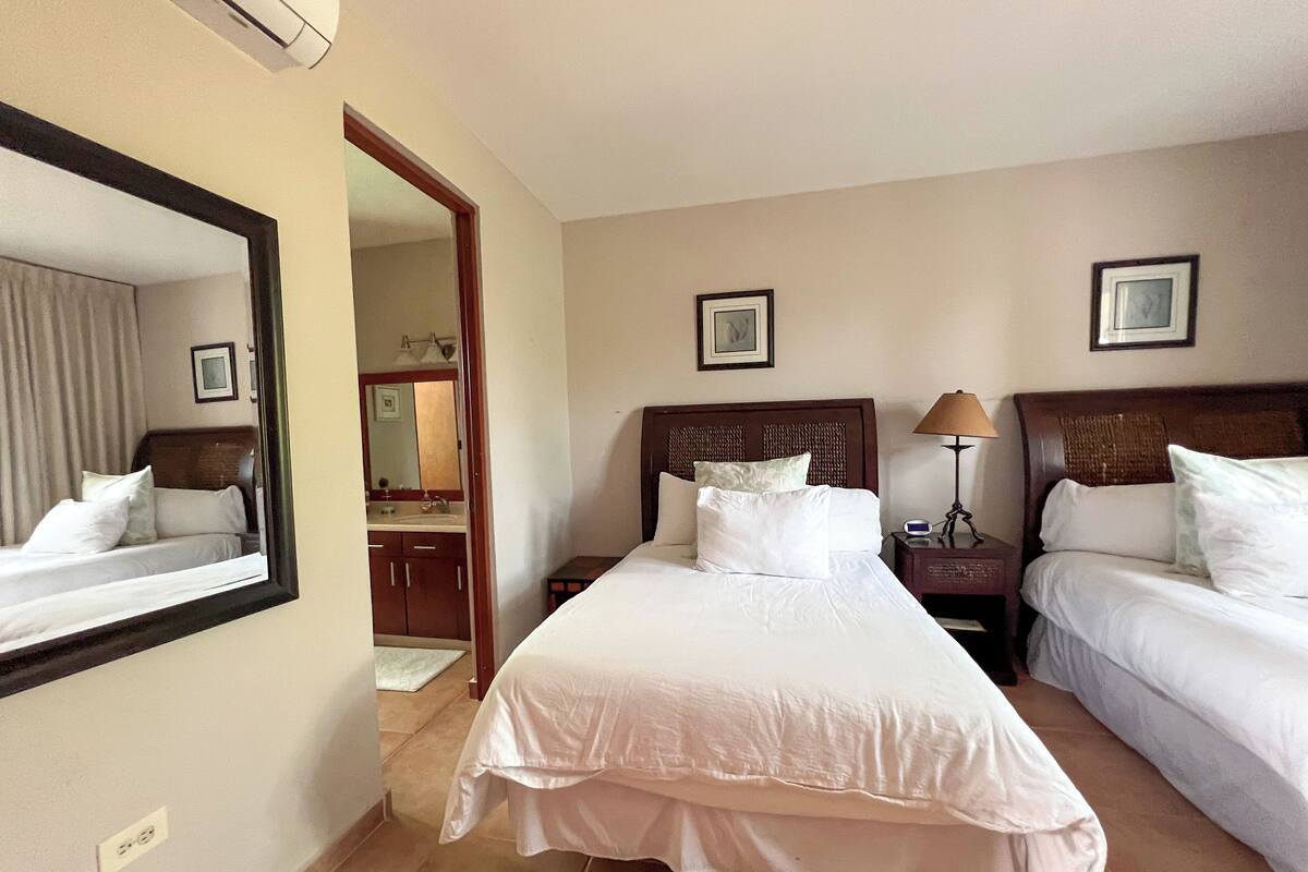 A hotel room with two single beds, a large mirror, a bedside table with a lamp, and a bathroom visible through an open door.