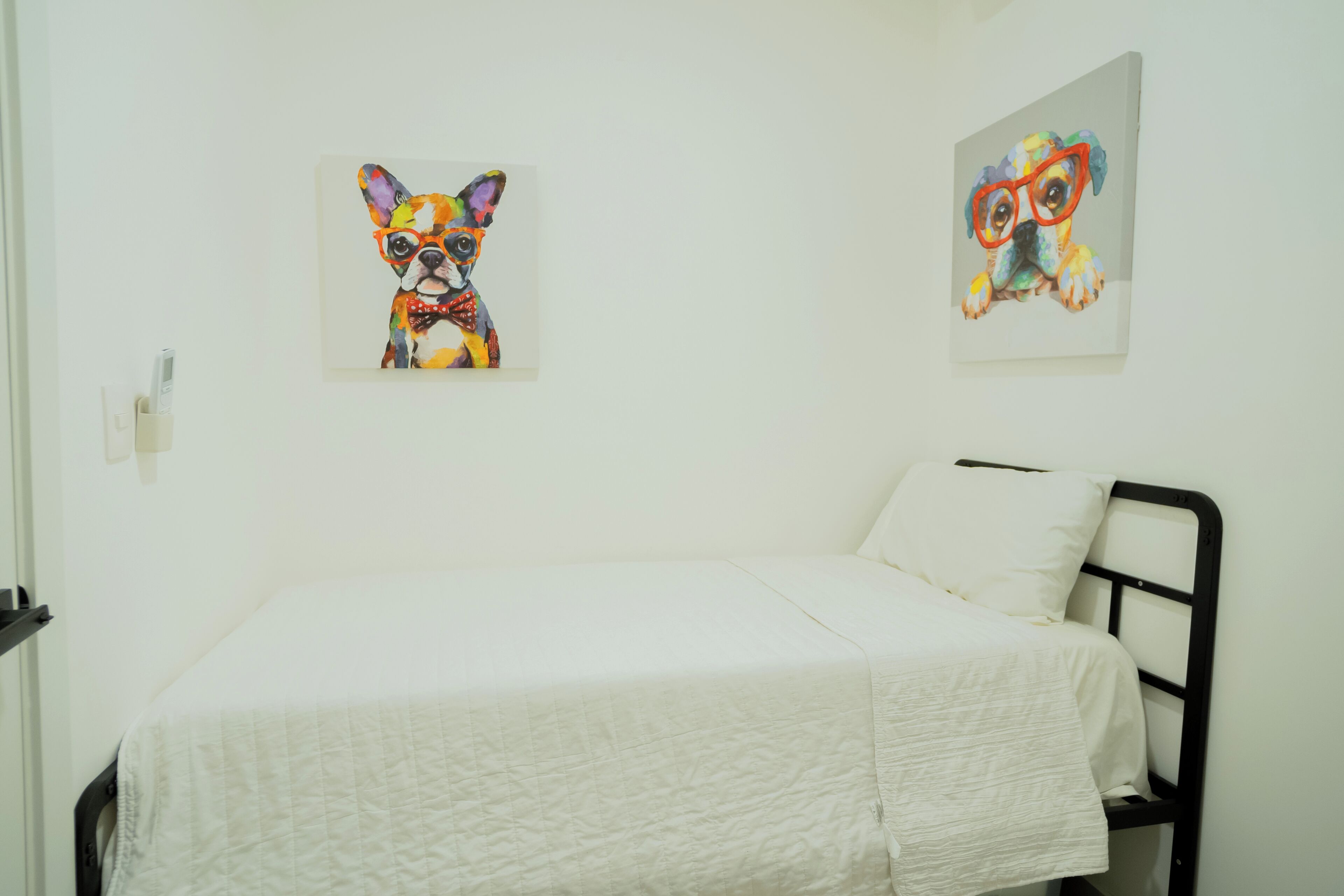 A single bed with a white quilted bedspread in a room with white walls. There are two framed artworks on the walls, one depicting a colorful dog and the other a dog with glasses.
