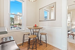 A room with a round table, three stools, a large mirror, and a view of buildings outside. - 2 BR Next To Lucas Oil - Walk to Conventions! (Indianapolis)