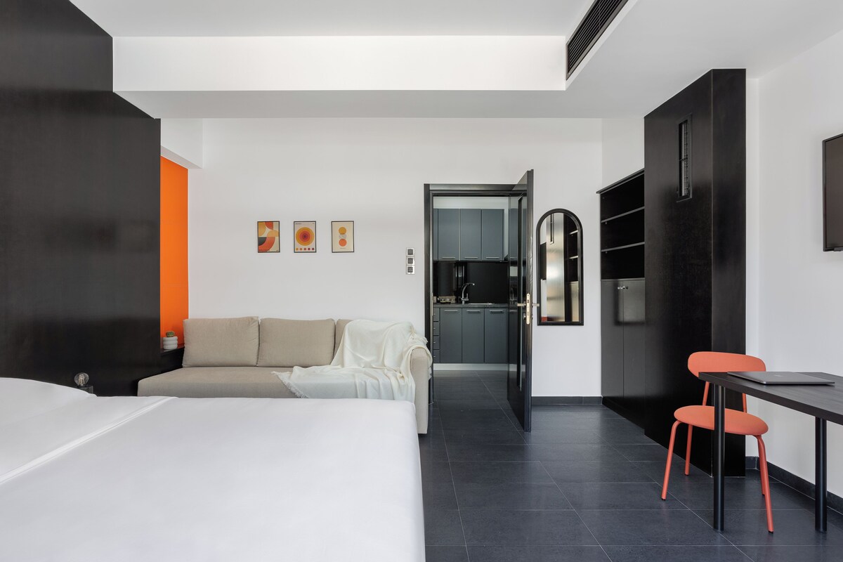 Una habitación de hotel moderna con cama, sofá, mesa de comedor con sillas, zona de cocina y obras de arte en las paredes.