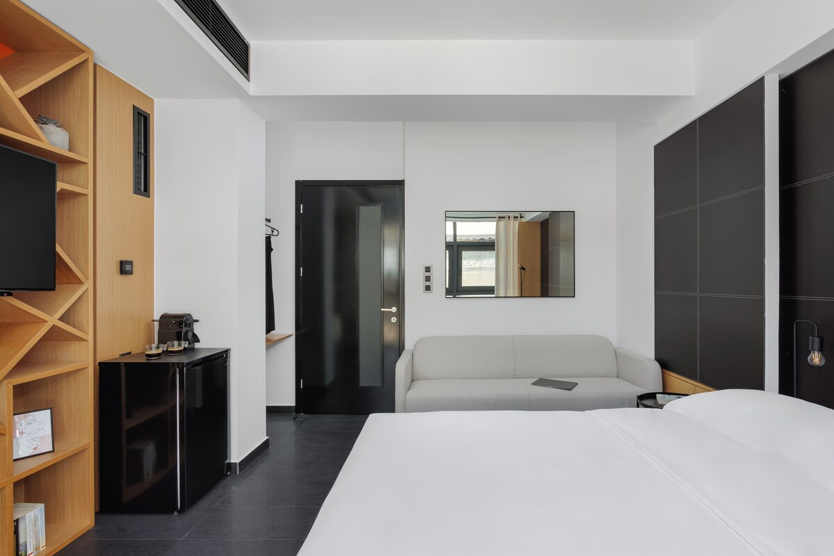 Una habitación de hotel moderna con una cama grande, un sofá, un televisor, una mesita y una estantería.