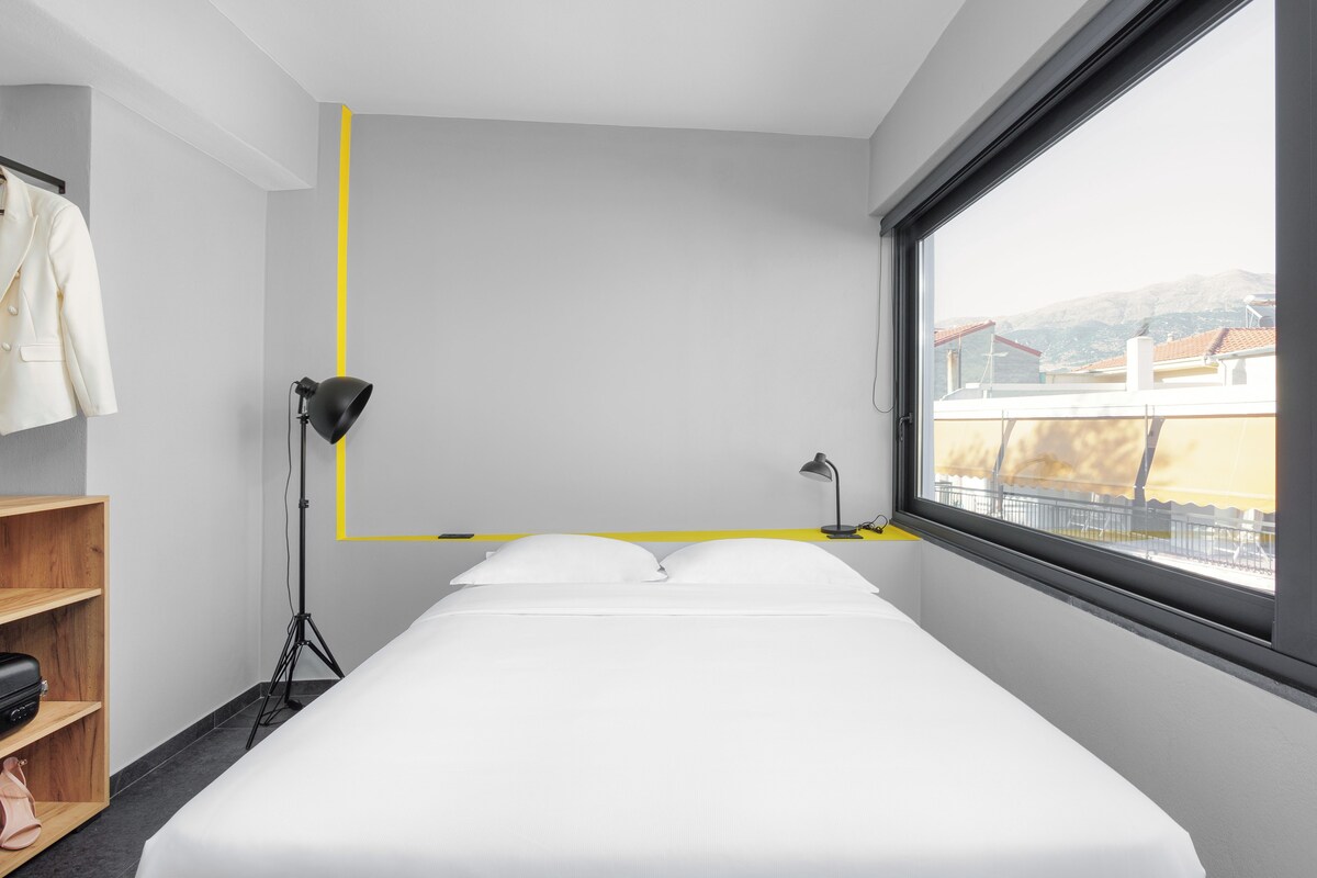 A modern hotel room with a large bed, a wardrobe, a desk, and a window with a view of buildings and hills.