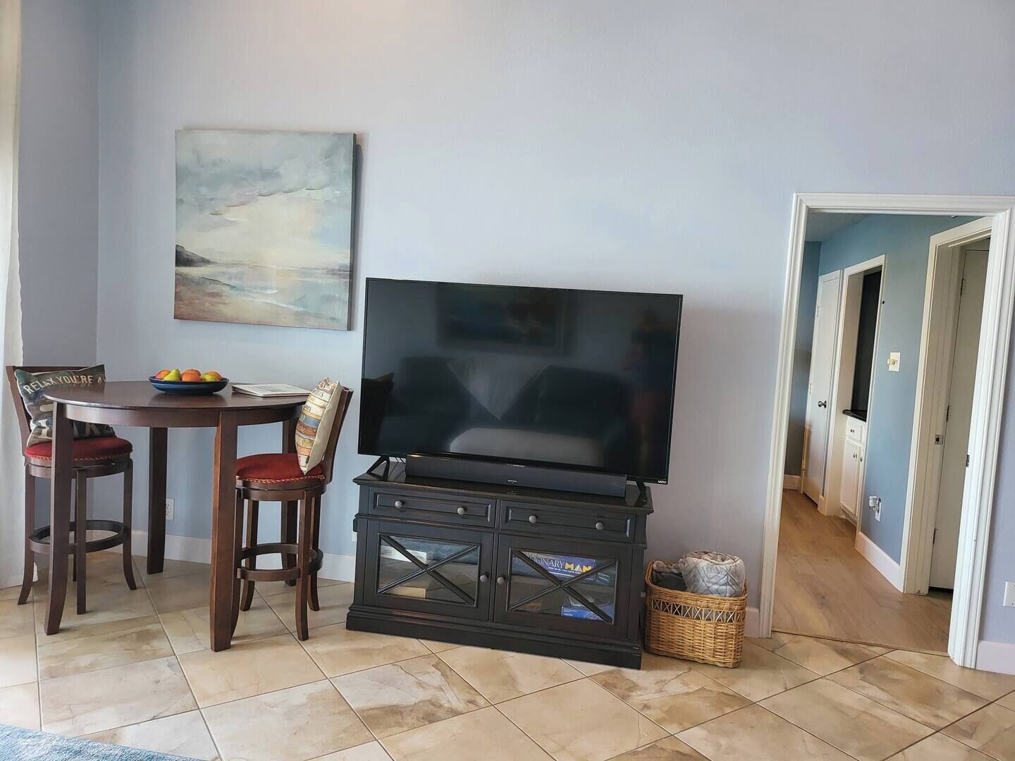 Ein Wohnzimmer mit Flachbildfernseher auf einem Standfuß, einem Esstisch mit Stühlen, einem Bild an der Wand und einem Flur, der in einen anderen Raum führt.