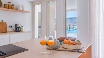 Une table dressée pour le petit-déjeuner, avec deux verres de jus d’orange, une théière, des croissants et un saladier de fruits, le tout placé devant une table offrant une vue sur une piscine et des montagnes.