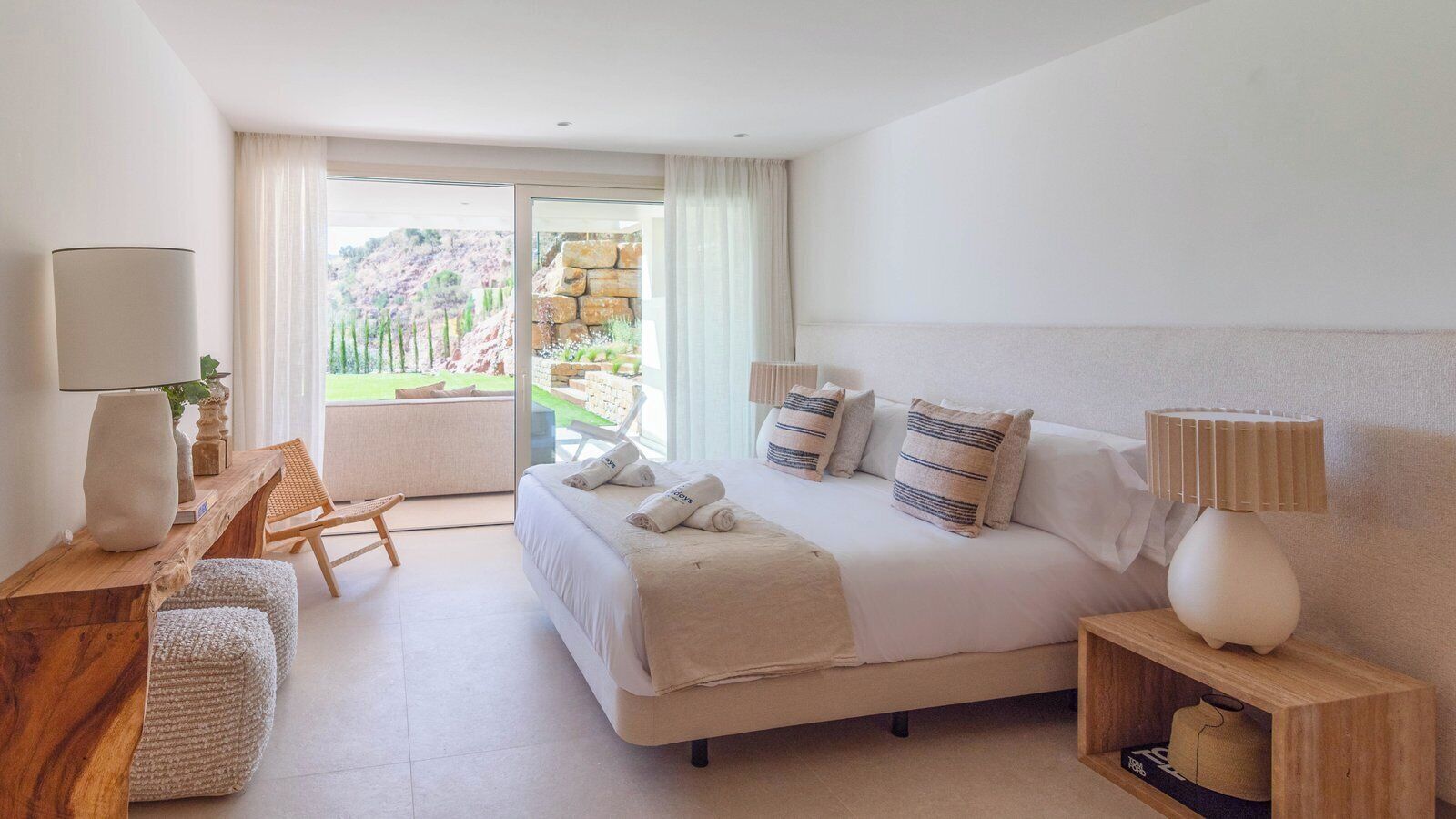 A modern hotel room with a large bed, a wooden desk, a chair, and a view of a garden through a glass door.
