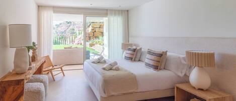 A modern hotel room with a large bed, a wooden desk, a chair, and a view of a garden through a glass door.